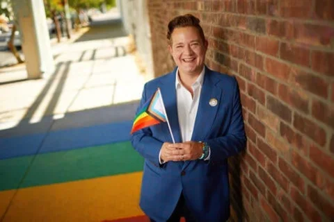 Man smiling with white shirt and blue jacket holding the LGBTQAI+ flag