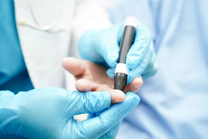 Gloved hands of a doctor use a lancet on a patient's finger to check blood sugar levels.
