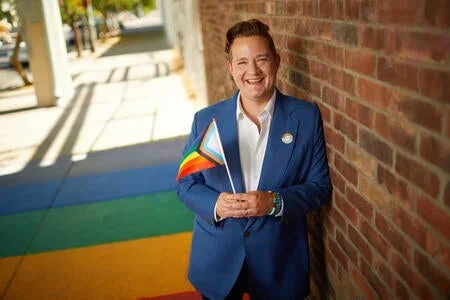 Man smiling with white shirt and blue jacket holding the LGBTQAI+ flag