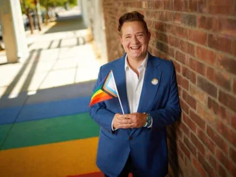 Man smiling with white shirt and blue jacket holding the LGBTQAI+ flag